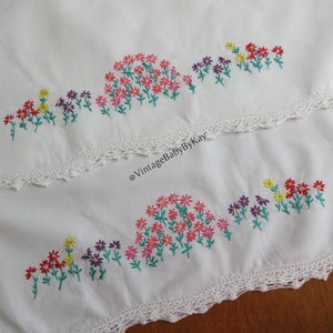 Pu&ograve; includere: Due pezzi di tessuto bianco con motivi floreali ricamati in rosa, rosso, giallo e viola con steli verdi. Ogni pezzo presenta un bordo decorativo in pizzo all'uncinetto bianco. Il testo @VintageBabyByKay &egrave; stampato al centro.