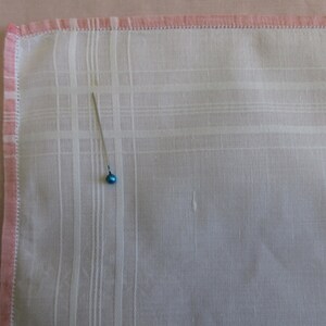May include: A white handkerchief with a pink border and a small blue pin in the center.