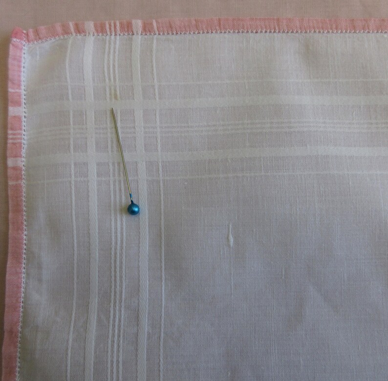 May include: A white handkerchief with a pink border and a small blue pin in the center.