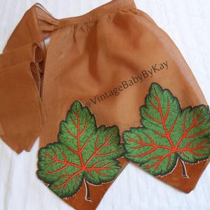 May include: A sheer brown apron featuring two large green leaf appliqués with prominent red veins. The apron has a gathered waist and long ties. The text @VintageBabyByKay is printed diagonally across the fabric in a dark brown font.