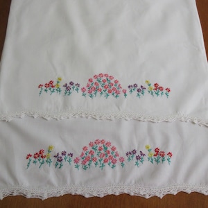 Pu&ograve; includere: Due pezzi di tessuto rettangolari bianchi con una fila orizzontale di fiori ricamati nei toni del rosa, rosso, giallo e viola con steli verdi. Ogni pezzo &egrave; rifinito con un delicato bordo in pizzo all'uncinetto bianco sul fondo.