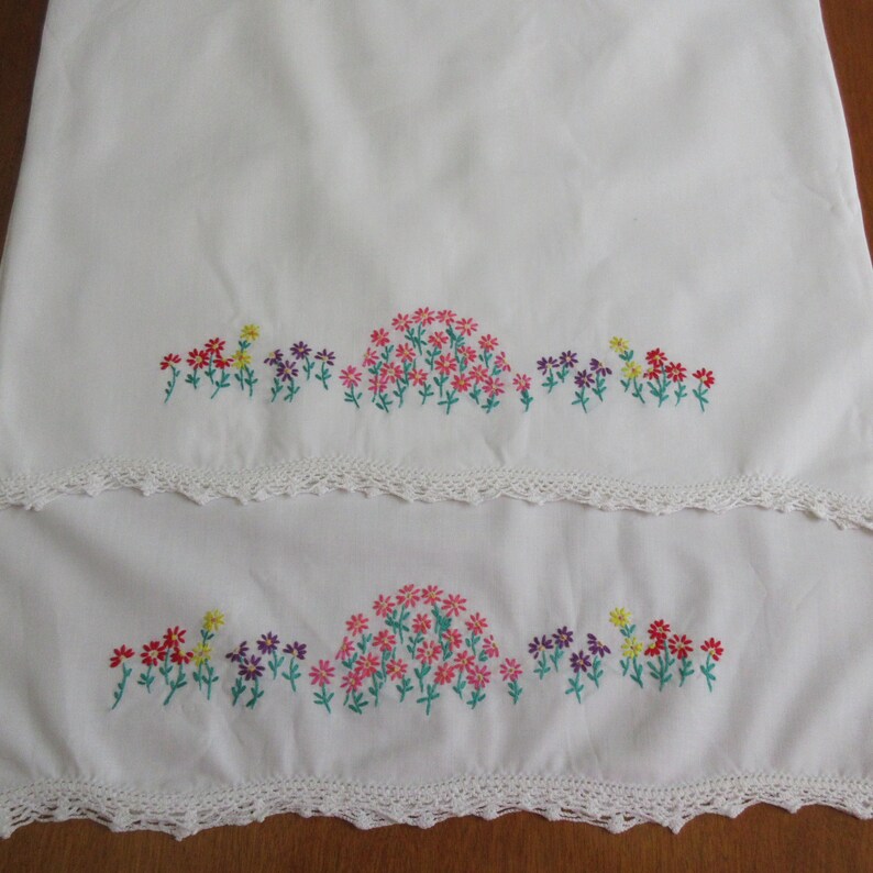 Pu&ograve; includere: Due pezzi di tessuto rettangolari bianchi con una fila orizzontale di fiori ricamati nei toni del rosa, rosso, giallo e viola con steli verdi. Ogni pezzo &egrave; rifinito con un delicato bordo in pizzo all'uncinetto bianco sul fondo.