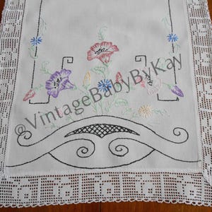 May include: White embroidered table runner with a floral design. The runner features a central embroidered bouquet of pink, purple, and yellow flowers, with black outlined decorative elements. The edges are trimmed with a white lace.