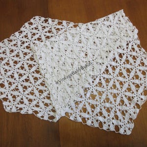 May include: Two white, open-weave crochet doilies with a floral pattern. The doilies are rectangular with scalloped edges. The doilies are displayed on a brown wooden surface. The text "@VintageBabyByKay" is visible on the doilies.