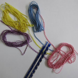 May include: A collection of colorful, wavy ribbons in yellow, blue, pink, and purple. A blue and silver sewing gauge is also present. The ribbons are arranged on a white surface, suggesting a craft or sewing project.