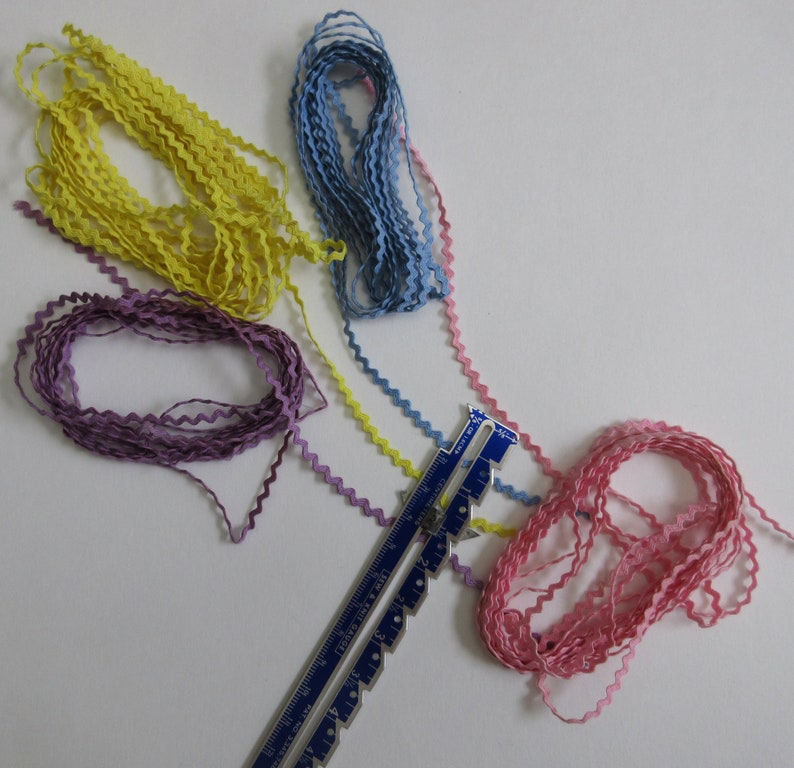May include: A collection of colorful, wavy ribbons in yellow, blue, pink, and purple. A blue and silver sewing gauge is also present. The ribbons are arranged on a white surface, suggesting a craft or sewing project.