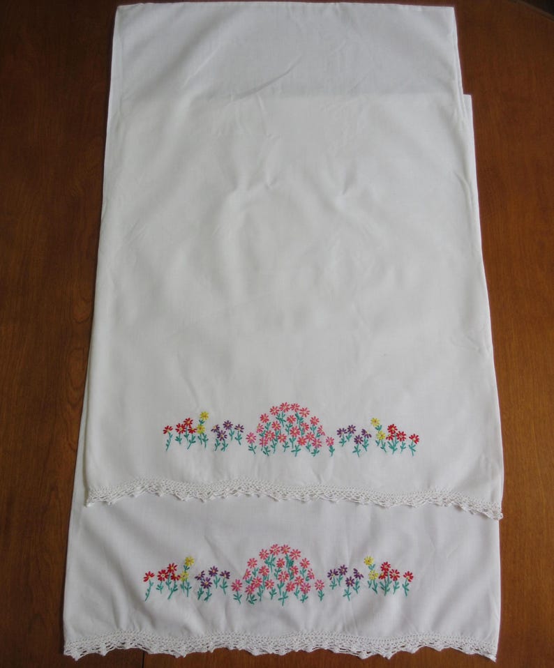 Pu&ograve; includere: Una coppia di federe rettangolari bianche con un motivo floreale ricamato a mano lungo il bordo inferiore. Il ricamo mostra piccoli fiori rossi, rosa, viola e gialli con steli verdi. Ogni federa presenta un bordo in pizzo bianco lavorato all'uncinetto.
