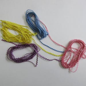 May include: A collection of colorful, wavy ribbons in shades of yellow, purple, blue, pink, and a mix of colors. The ribbons are arranged on a white surface, with each color in a separate pile or strand. The edges of the ribbons have a zig-zag pattern.