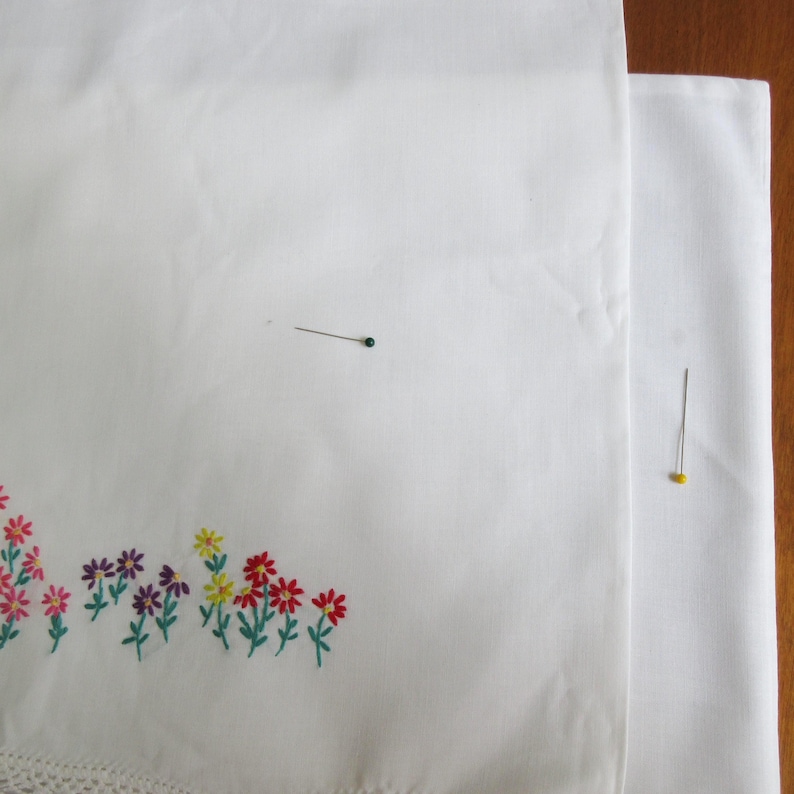 Pu&ograve; includere: Un pezzo di tessuto bianco con un motivo floreale ricamato colorato lungo il bordo inferiore, che mostra piccoli fiori rossi, viola e gialli con steli verdi. Due spilli da cucito con teste colorate sono posizionati sul tessuto.