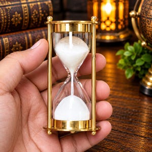 Puede incluir: Un reloj de arena de latón con un bulbo de vidrio transparente lleno de arena blanca. El marco consta de dos placas circulares de latón conectadas por cuatro varillas verticales delgadas de latón, con pequeñas esferas decorativas.