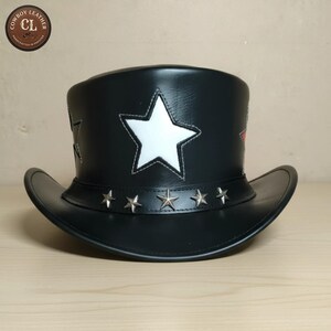 May include: A black leather top hat featuring a large white star appliqué on the front and a band decorated with five small silver-toned metal star studs. The hat has a curved brim and includes a circular logo in the corner reading COWBOY LEATHER CL.