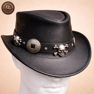 May include: A black leather cowboy hat featuring a wide brim and a crown with two ventilation eyelets on the side. The hat band is adorned with silver-toned metal skull emblems, crossbones, and a central circular medallion with a square cutout.