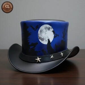 May include: A black top hat featuring a blue night sky scene with a large white full moon, a howling wolf silhouette, and flying bats. The hat includes a black leather band adorned with four silver-toned metal star-shaped studs.