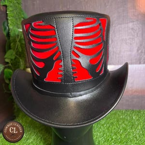 May include: A black leather top hat featuring a prominent cutout design of a human rib cage on the front. The cutout reveals a vibrant red fabric layer underneath the black leather structure, creating a high-contrast skeletal appearance.