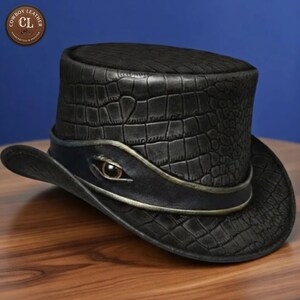 May include: A black top hat with a textured crocodile skin pattern. The hat features a smooth black leather band wrapped around the base, accented with a single realistic eye detail centred on the front. A circular logo reads Cowboy Leather CL.