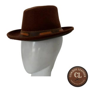 Pode incluir: Um chapéu fedora de feltro castanho escuro com aba larga e uma faixa de couro decorativa com atacadores de couro castanho claro. O chapéu é exibido numa cabeça de manequim branca. Um logótipo circular indica Cowboy Leather CL Handcrafted in Pakistan.