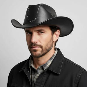 Könnte beinhalten: Ein schwarzer Cowboyhut mit einer strukturierten Krone und weißen Ziernahtdetails am Vorderteil. Der Hut hat eine breite, geschwungene Krempe und kleine Belüftungsösen aus Metall an den Seiten, gefertigt aus einem glatten, lederartigen Material.
