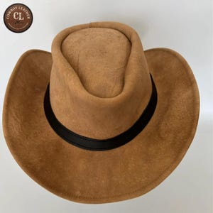 Könnte beinhalten: Ein beigefarbener Wildleder-Cowboyhut mit breiter Krempe und einem glatten schwarzen Lederband um die Krone. Ein kreisförmiges Logo in der oberen linken Ecke zeigt den Text Cowboy Leather CL Handcrafted in Pakistan mit einem kleinen Hut-Symbol.