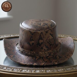 May include: A brown and black snakeskin patterned leather hat with a flat top and a wide brim. The hat features a decorative braided leather band around the base of the crown. A circular logo in the top left corner reads Cowboy Leather CL.