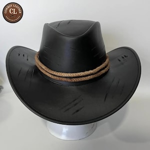Peut inclure: Un chapeau de cowboy en cuir noir avec une finition vieillie présentant des marques de griffures intentionnelles sur la calotte et les bords. Le chapeau est orné d'une bande en corde beige à double brin autour de la base. Un logo circulaire en haut à gauche indique COWBOY LEATHER CL HANDCRAFTED IN PAKISTAN.