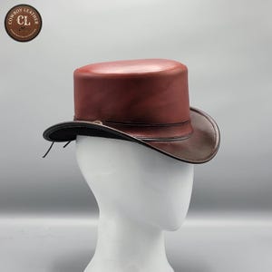 Puede incluir: Un sombrero de copa hecho de cuero liso de color rojo oscuro con una banda de cuero marrón oscuro que rodea la base de la copa. El ala está ligeramente curvada y el sombrero presenta un pequeño detalle metálico en la banda. Un logo dice COWBOY LEATHER.