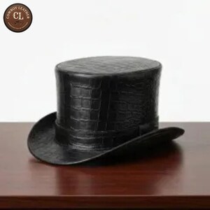 Può includere: Un cappello a cilindro nero con un motivo testurizzato in pelle di coccodrillo. Il cappello presenta una corona cilindrica alta e una tesa curva. Un logo circolare nell'angolo in alto a sinistra mostra il testo Cowboy Leather CL Handcrafted in Pakistan.