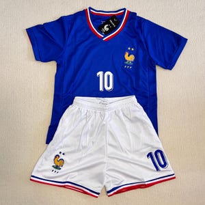 Puede incluir: Una camiseta azul de manga corta con cuello en V con ribetes rojos y blancos, el número 10 en blanco y un emblema de gallo con dos estrellas. Se combina con pantalones cortos blancos con el número 10, un emblema de gallo y ribetes tricolores.