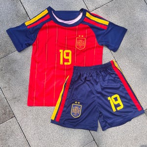 May include: A red and blue soccer jersey and shorts set featuring yellow accents. The jersey has the number 19 in yellow, a crest with two stars, and thin vertical lines. The blue shorts feature the number 19 in yellow and a matching crest.