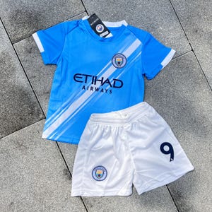 May include: A light blue short-sleeved jersey with white diagonal stripes, the Etihad Airways logo, and the Manchester City crest. Paired with white shorts featuring the number 9 in dark blue and the Manchester City crest on the right leg.