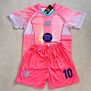 Pode incluir: Um conjunto de futebol rosa composto por uma camisola e calções a condizer. A camisola apresenta um decote em V, o emblema do FC Barcelona, um logótipo Spotify e uma etiqueta. Os calções exibem o emblema do FC Barcelona e o número 10 em azul.
