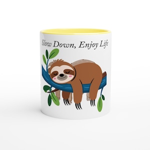 Peut inclure: Un mug en céramique blanc avec un bord intérieur jaune, illustré d'un paresseux brun allongé sur une branche d'arbre bleue avec des feuilles vertes. Le texte "Slow Down, Enjoy Life" est imprimé en noir au-dessus du paresseux sur le mug.
