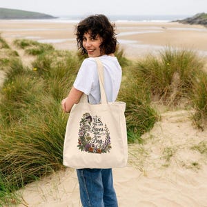 May include: A cream-colored canvas tote bag featuring a colorful botanical illustration of various flowers and foliage surrounding the black text that reads, YES, THERE ARE LEAVES IN MY BAG. The bag has two shoulder straps and a rectangular shape.