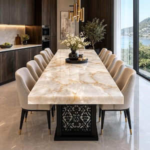 Op de afbeelding: Een lange rechthoekige eettafel met een gepolijst wit en goud marmeren blad en een zwart geometrisch roosteronderstel. De tafel is omringd door twaalf gestoffeerde beige stoelen met zwarte poten en goudkleurige metalen uiteinden bij de voeten.