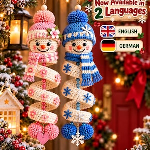 May include: Two crocheted snowman ornaments hanging vertically. The left snowman wears a pink hat and scarf with flower accents. The right snowman wears a blue hat and scarf with snowflake accents. Text reads: Now Available in 2 Languages.