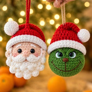 Op de afbeelding: Twee gehaakte kerstornamenten met een kerstmangezicht met witte baard en rode muts, en een groen karaktergezicht met rode muts. Beide ornamenten hebben zwarte plastic ogen en zijn voorzien van rode en bruine touwtjes om ze op te hangen.