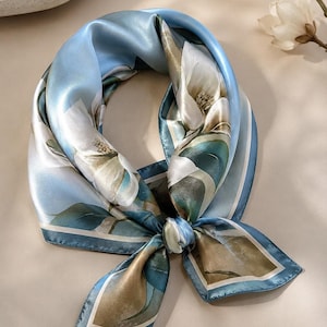 May include: A square silk scarf featuring a light blue background with a large white magnolia flower print and dark green foliage. The scarf is tied in a knot, displaying a smooth, reflective surface with a thin white border along the edges.