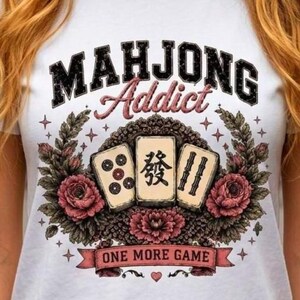 May include: A white short-sleeved t-shirt featuring a graphic print with the text "MAHJONG Addict" and "ONE MORE GAME". The design includes three mahjong tiles surrounded by dark green leaves, pink roses, and small star accents in a vintage style.