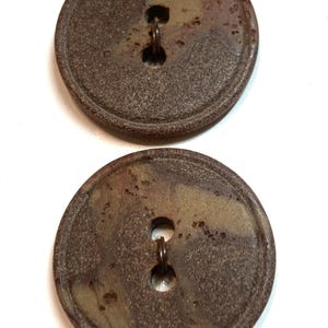May include: Two round, brown buttons with a mottled appearance. Each button has a metal shank for sewing. The buttons have a rustic, vintage aesthetic, with a slightly textured surface and a diameter of approximately 1.25 inches.
