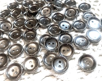 May include: A collection of shiny, silver-colored metal buttons with two holes each. The buttons are round and slightly domed, scattered on a white, textured surface. The buttons are likely for sewing or crafting projects.