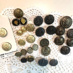 May include: A collection of vintage buttons in various sizes and designs. The buttons are in shades of gold, black, and bronze, featuring crests, emblems, and ornate details. Some buttons are attached to a tag that reads "JHB International BUTTONS".