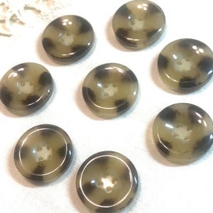 May include: A collection of eight round buttons with a tortoiseshell pattern. The buttons are primarily tan with dark brown spots and a four-hole design. They are arranged on a white, textured surface.