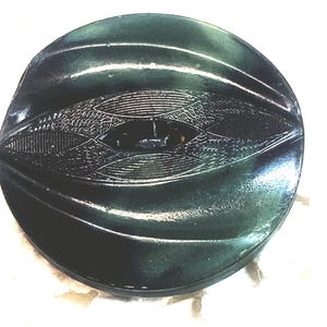 May include: A dark green, round button with an eye-shaped, textured design in the center. The button has a glossy finish and a slightly raised, ridged pattern around the edges. The central design features intricate detailing.