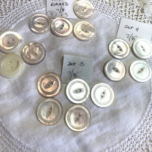May include: A collection of white pearl buttons with two holes. The buttons are arranged in a circle on a white fabric background. The buttons are different sizes, with some being 7/8 inches in diameter. There are two sets of buttons, one labeled "Set 4 7/8" and the other labeled "Set 5 7/8".