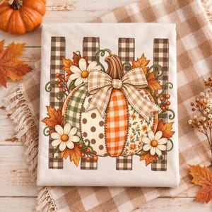 May include: A white t-shirt featuring a central graphic of a pumpkin composed of various patterned segments including green plaid, brown polka dots, orange gingham, and floral prints. The pumpkin is adorned with a beige gingham bow, white daisies, and autumn leaves.