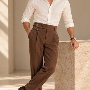 Puede incluir: Una camisa de manga larga abotonada de color crema combinada con pantalones de pana marrones de cintura alta con detalle de lengüeta lateral de doble botón, frente plisado y dobladillos con vuelta, junto con mocasines de cuero marrón.