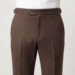 Könnte beinhalten: Braune Hose mit dezentem Fischgrätenmuster, hohem Bund mit einem einzelnen mittigen Knopfverschluss und seitlichen Verstelllaschen. Die Hose wird mit einem knackig weißen Hemd kombiniert, das in den Bund gesteckt ist und eine klare Silhouette bildet.