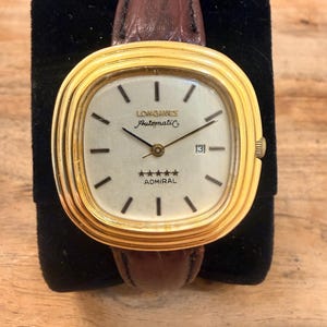 May include: A Longines Admiral automatic wristwatch with a gold-tone cushion-shaped case and a white dial featuring black hour markers, a date window at the three o'clock position, five stars, and a brown textured leather strap with a buckle.