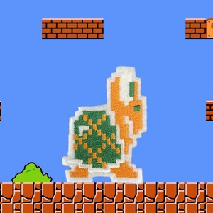 Może przedstawiać: Zielona i pomarańczowa pikselowa postać żółwia z klasycznej gry wideo. Żółw stoi na platformie z cegieł z zielonym krzewem przed nim. Nad platformą znajdują się bloki z pytajnikami.