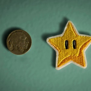 May include: A yellow star-shaped patch with a black outline and two black eyes. The patch is embroidered and has a white border.