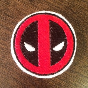 May include: Red and black embroidered patch with a white outline, featuring the Deadpool logo. The logo is a stylized red mask with black eyes and a white outline.